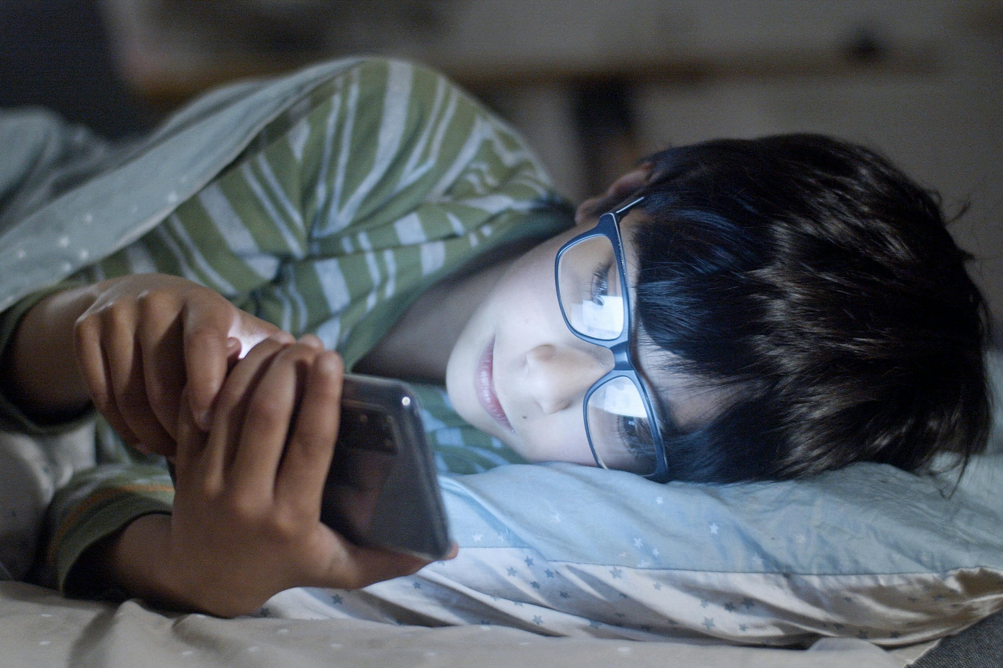 Child wearing glasses using smartphone in bed – late night screen use and blue light impact